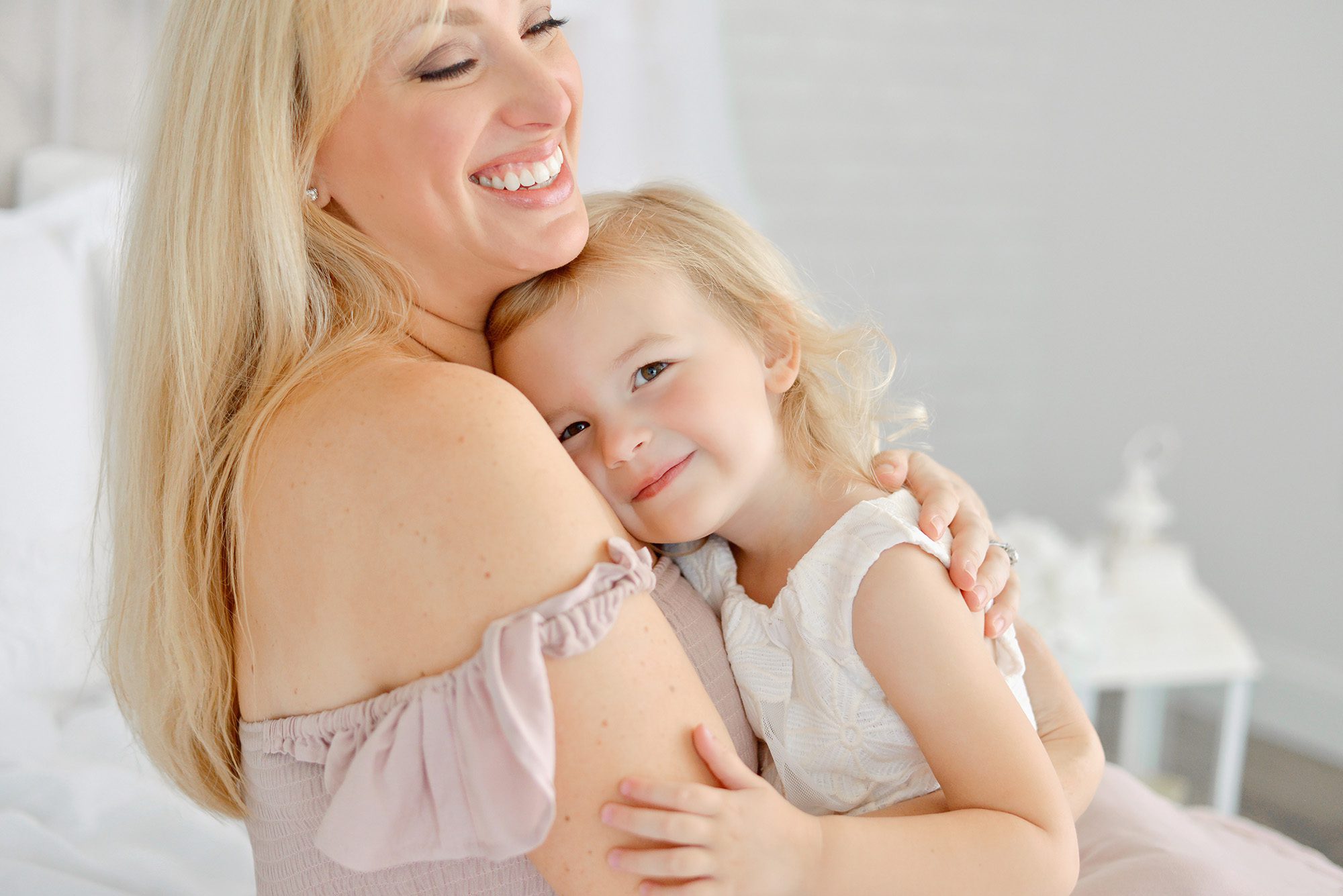 Mom and daughter happily snuggling and giggling on a white bed in a studio for a portrait session