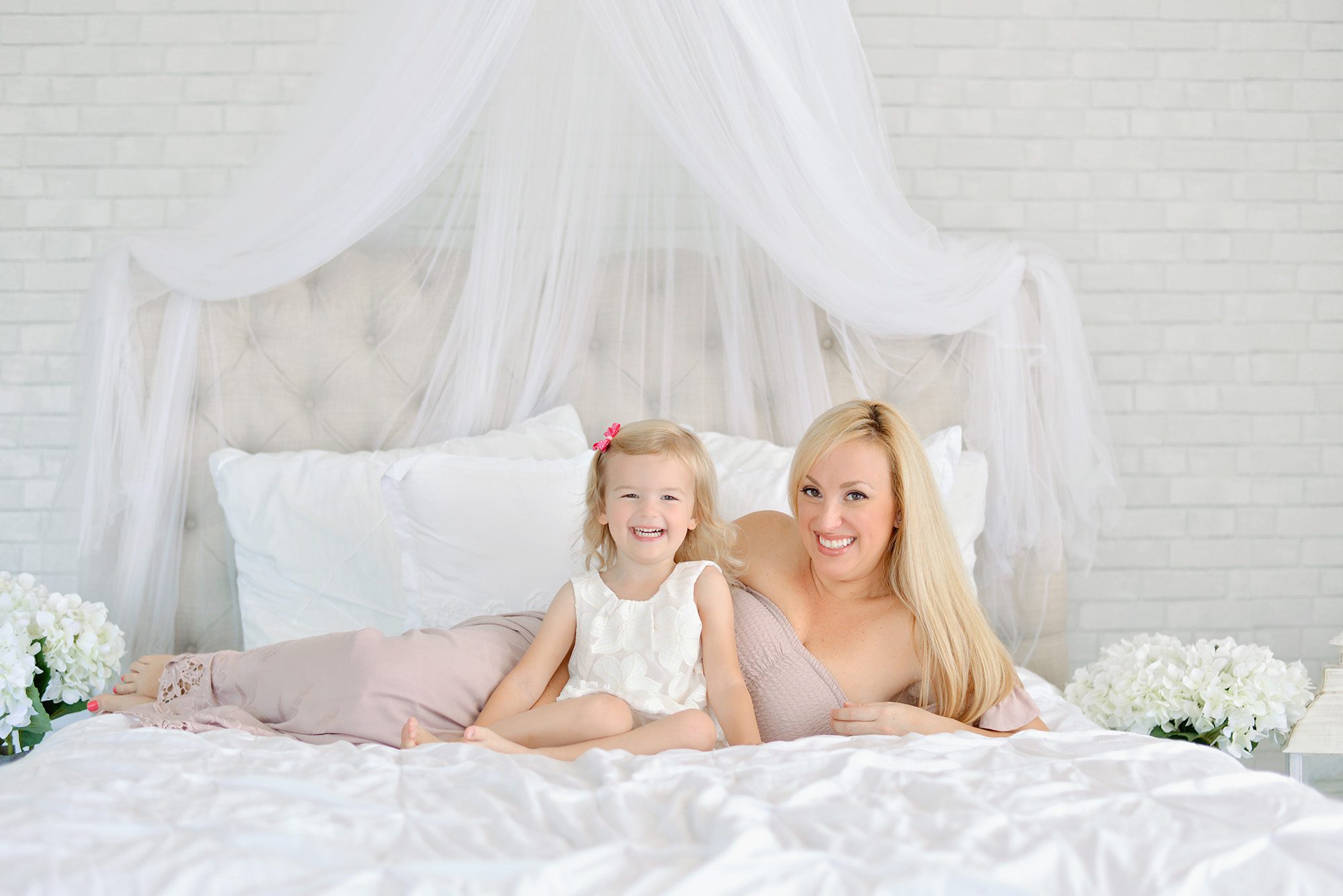 Mom and daughter happily snuggling and giggling on a white bed in a studio for a portrait session