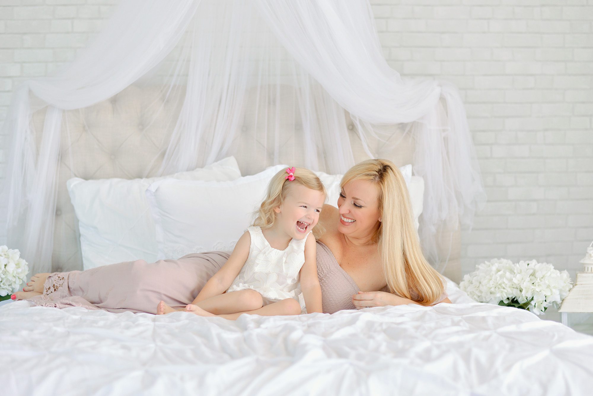 Mom and daughter happily snuggling and giggling on a white bed in a studio for a portrait session