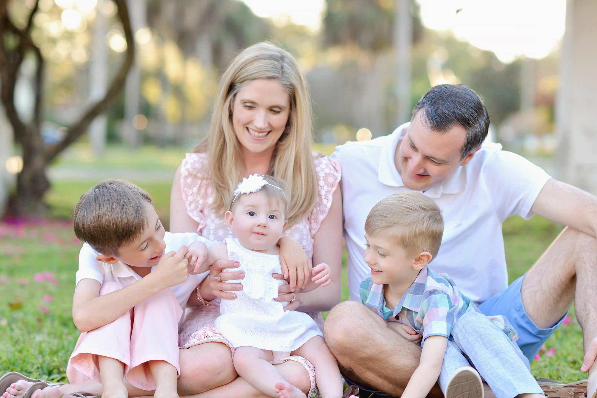 A family of five with 2 boys and a baby girl being photographed in a park with pink flowers and a white pergola in St Petersburg Florida.