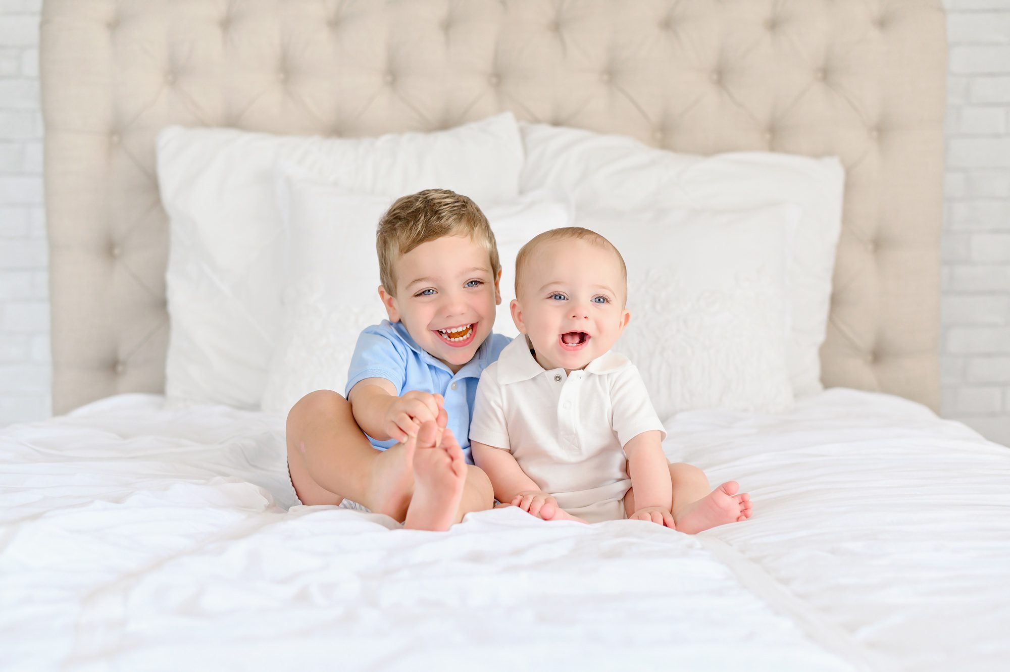 An adorable blue eyed baby boy gets his 6 month portraits done on a white bed in a studio in Tampa, FL and his older brother jumps in for a few.