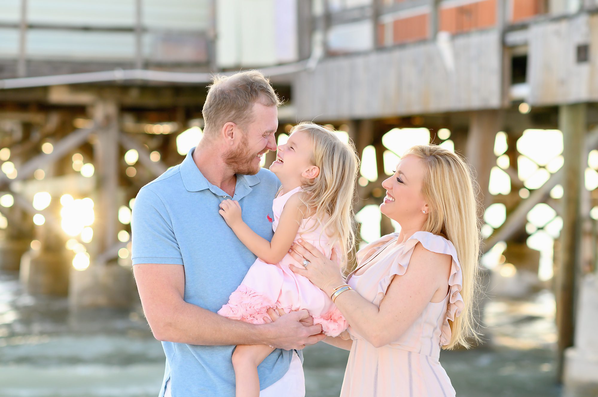 A mom and dad take family beach portraits with their young blonde haired daughter at sunset in front of Redington Pier in Redington Shores, Florida. 
