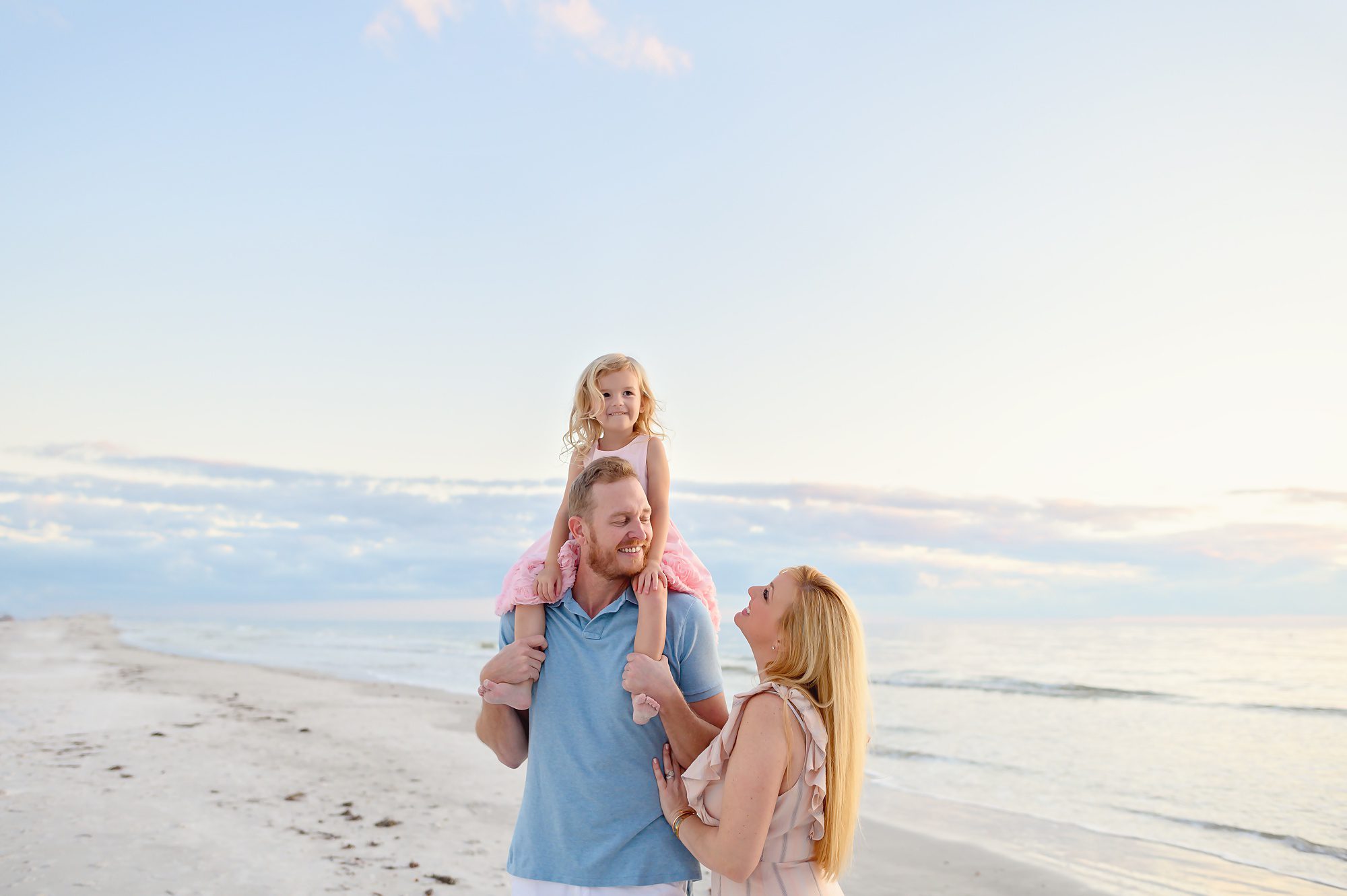 A mom and dad take family beach portraits with their young blonde haired daughter at sunset in front of Redington Pier in Redington Shores, Florida. 
