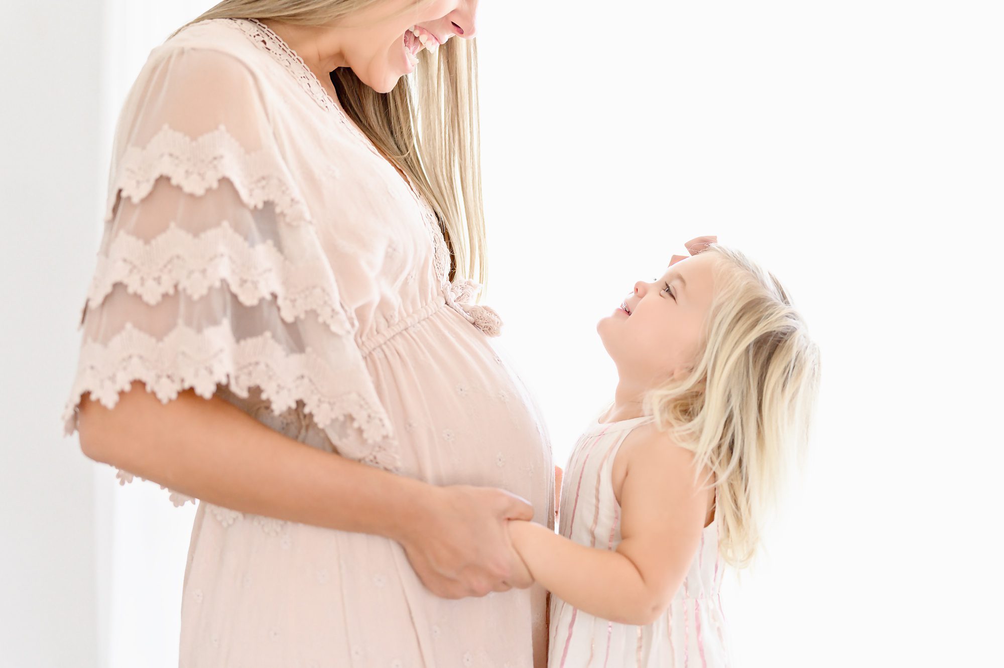 A young mom and her beautiful blond toddler do mommy and me maternity portraits in a bright white studio.