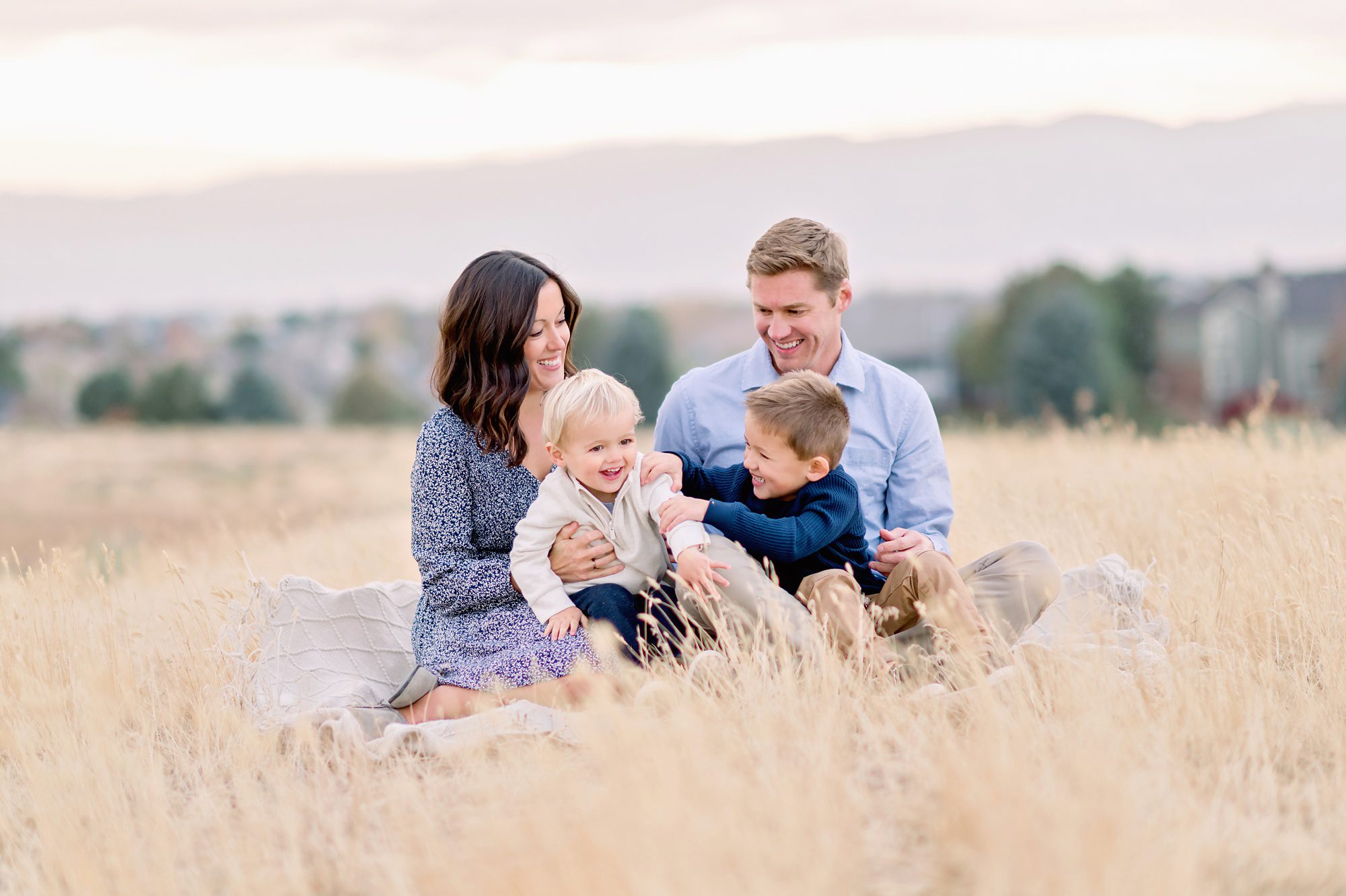 A family of 4 with 2 young boys get family photos taken in a field in Highlands Ranch, CO with a beautiful sunset and mountain backdrop.
